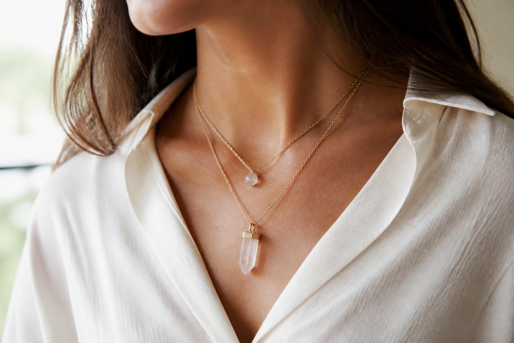Close-up of a woman's neck and collarbone wearing a layered crystal pendant necklace with a simple blouse, natural daylight, soft neutral background