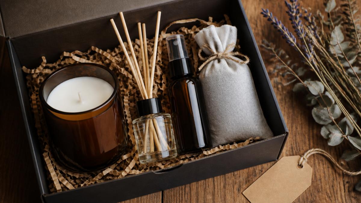 A flat-lay photograph of an open home fragrance gift box on a wooden table, showing a hand-poured candle in amber glass, a small reed diffuser with natural reeds, a room spray bottle, and a wrapped sachet, with dried botanicals and a handwritten gift tag beside it
