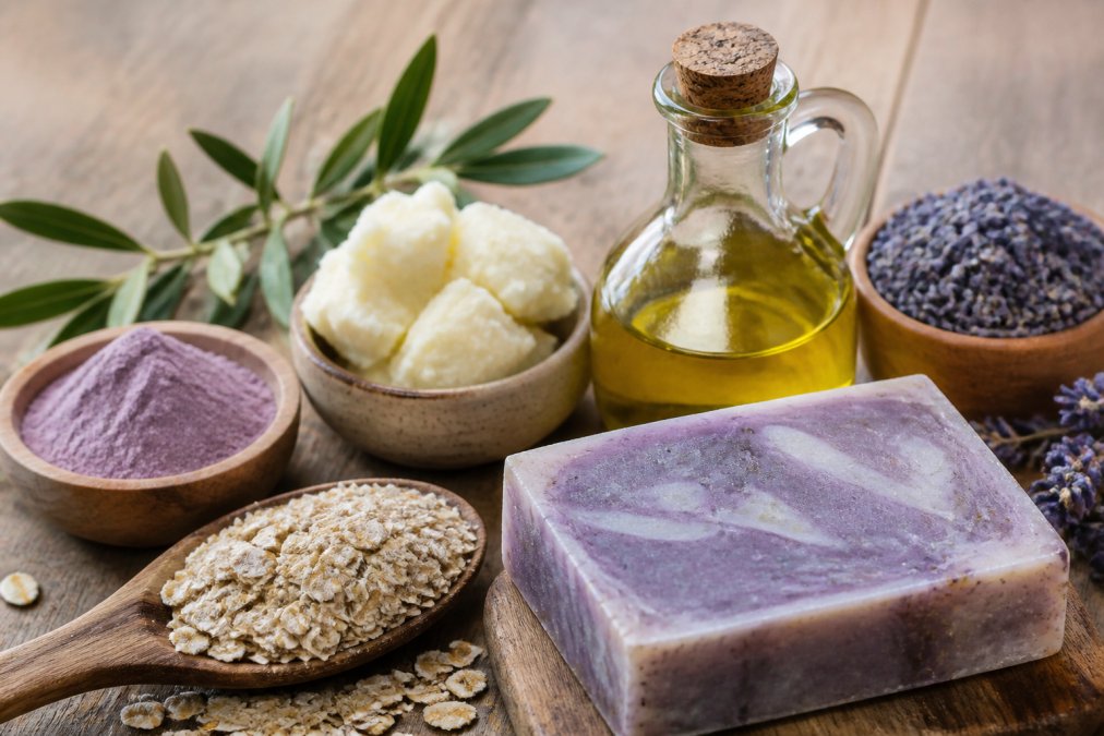 Close-up of natural soap ingredients arranged on a wooden table: cold-pressed olive oil in a glass bottle, raw shea butter, dried lavender, colloidal oats, and purple clay, next to a single finished artisanal soap bar with visible texture