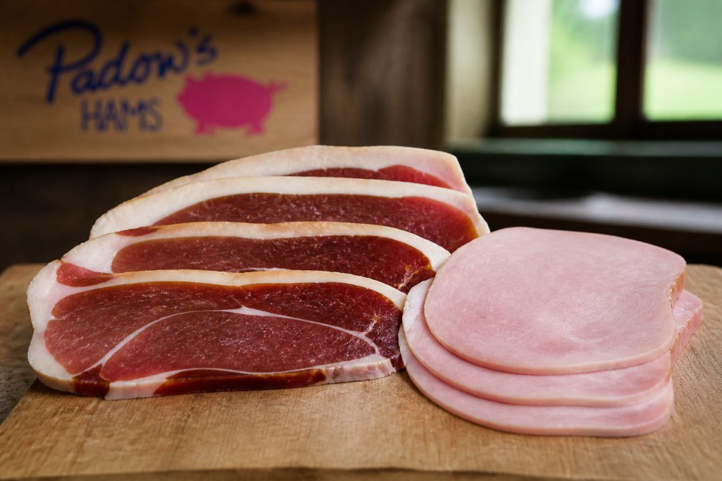 Close-up of a sliced artisanal country ham on a wooden cutting board showing deep mahogany color, visible marbling, and a thick fat cap, alongside a pale, uniform slice of mass-produced supermarket ham for visual contrast