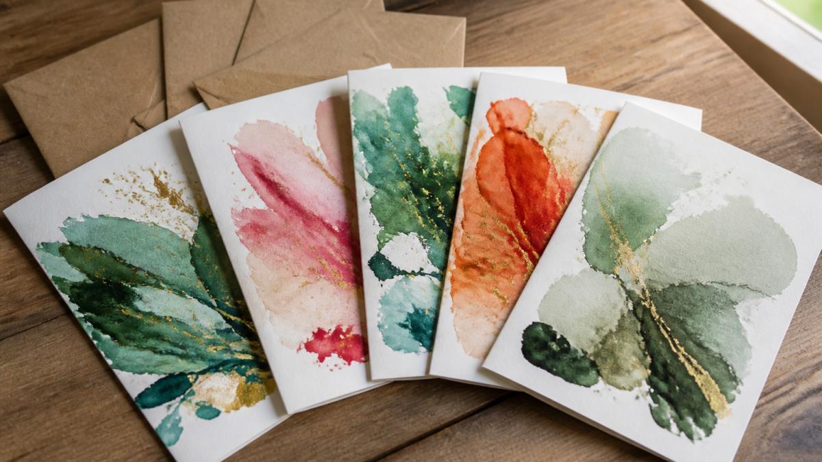 Close-up of five fine-art greeting cards fanned across a wooden surface, showing vibrant watercolour brushstrokes in greens, reds, and golds, with FSC kraft envelopes alongside and soft natural window lighting