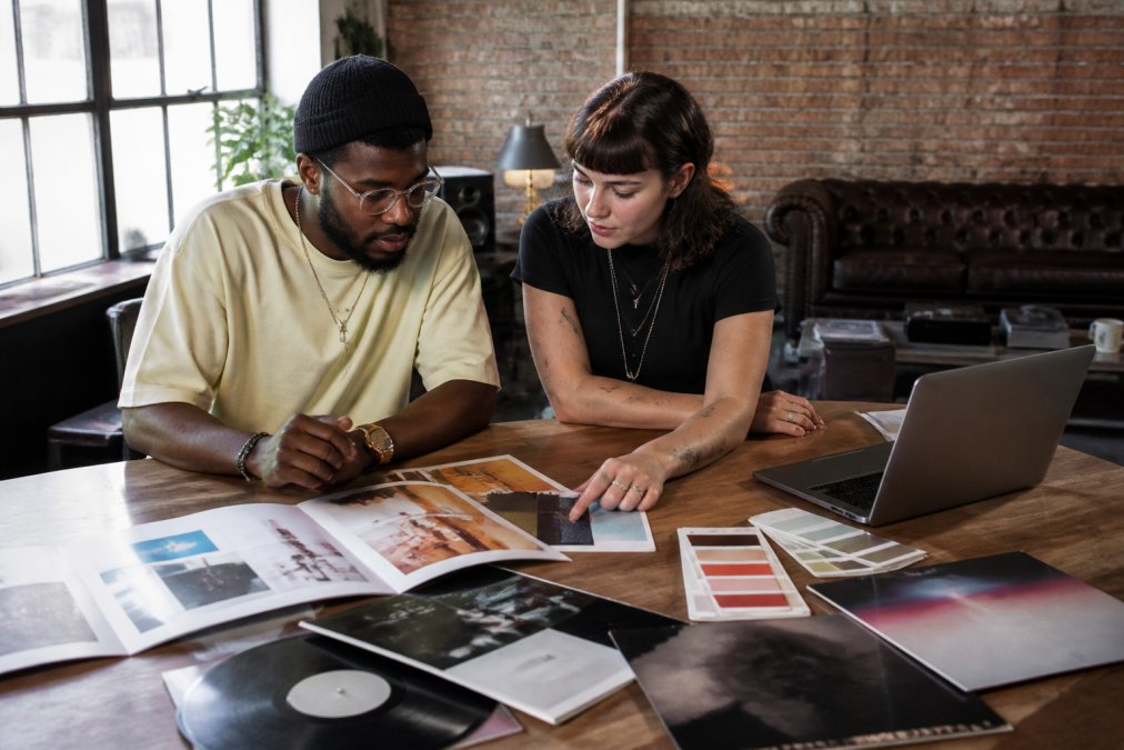 Independent record label manager and emerging artist reviewing brand identity mood boards and visual concepts together on a desk with laptops, color swatches, and album artwork mockups in a creative studio space with natural daylight