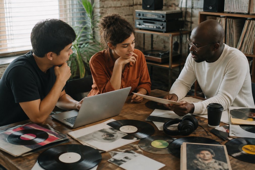 Small independent music label team of three people collaborating around a desk covered with vinyl records, laptop displaying streaming analytics, headphones, and artist promotional materials in a compact creative office with vintage equipment visible in background
