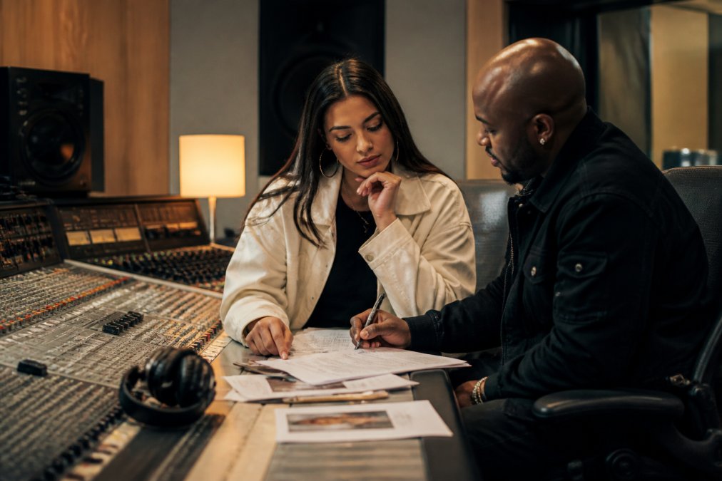 Music manager and artist in professional discussion at a studio mixing console with contracts and strategy notes spread on desk, natural studio lighting