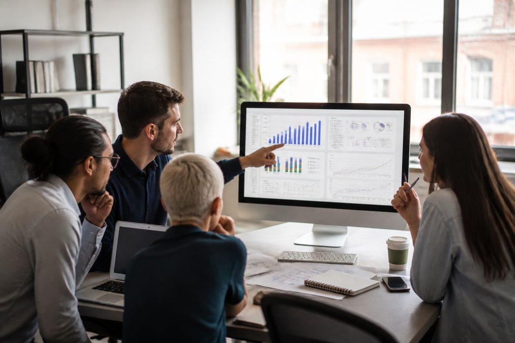Team of label executives collaborating around a desk, examining streaming and royalty performance metrics on a computer screen in a bright modern office with natural window lighting