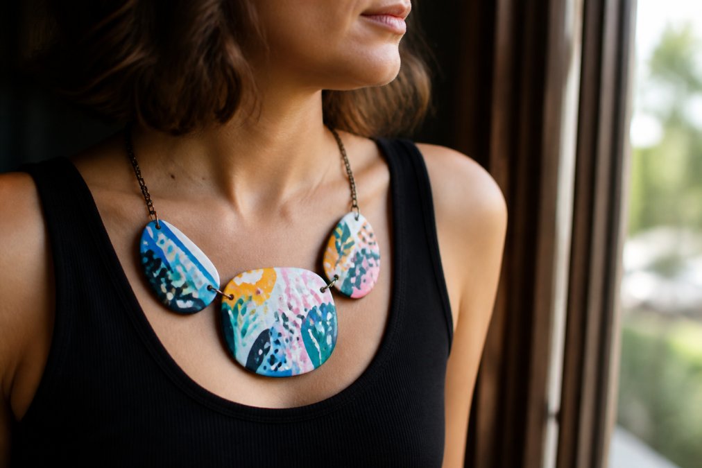 Close-up photograph of a person wearing a bold, hand-painted statement necklace over a simple black top, soft natural window light, shallow depth of field