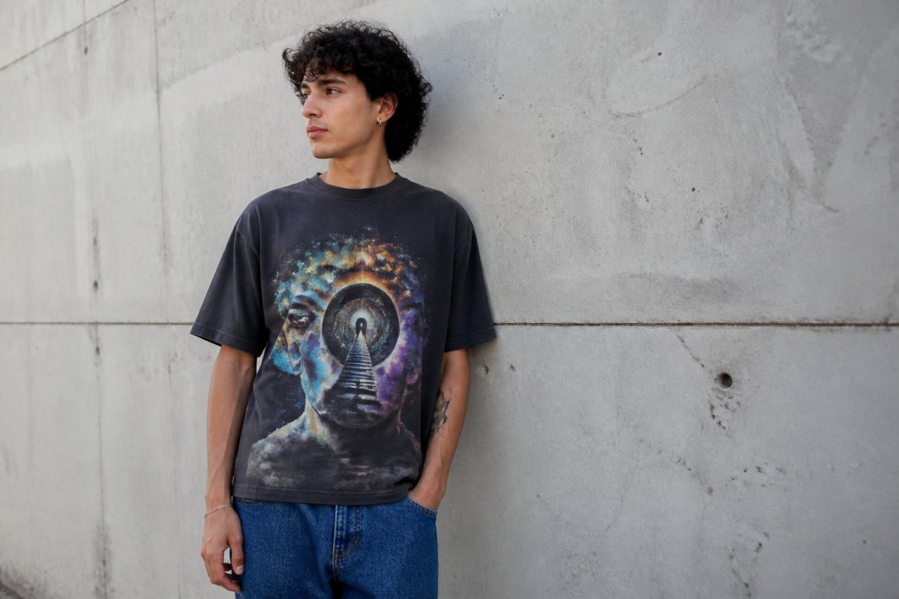 A young adult in their twenties wearing a bold, colorful artistic graphic tee featuring a surrealist design, standing against a plain concrete urban wall in soft natural daylight