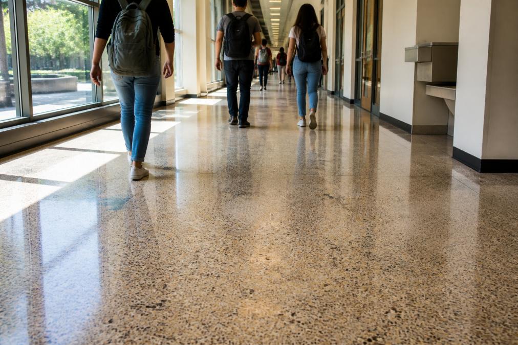 A wide, sunlit university corridor with students walking between classes, backpacks slung over shoulders, the polished flooring reflecting daylight and showing light surface wear in the main traffic path