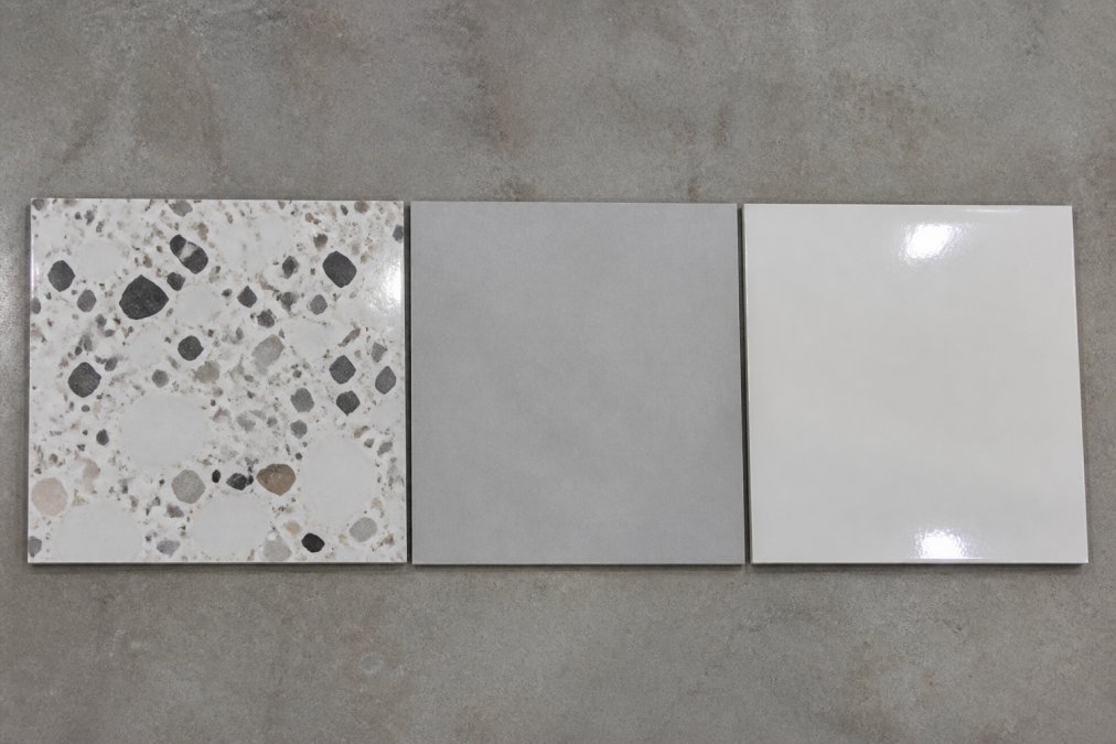Side-by-side view of three distinct flooring samples on a neutral backdrop: a polished terrazzo surface with large marble chips, a matte porcelain tile, and a glazed ceramic tile, lit by overhead track lighting