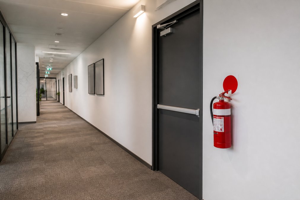 Wide view of a commercial corridor showing a fire extinguisher mounted on the wall to the side of an exit door, with clear space around it and an unobstructed pathway visible, demonstrating proper accessibility and egress compliance