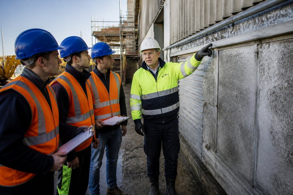 A construction site supervisor in high-visibility clothing pointing out potential asbestos-containing materials to a small group of tradespeople during a site induction, with clipboards and safety helmets visible