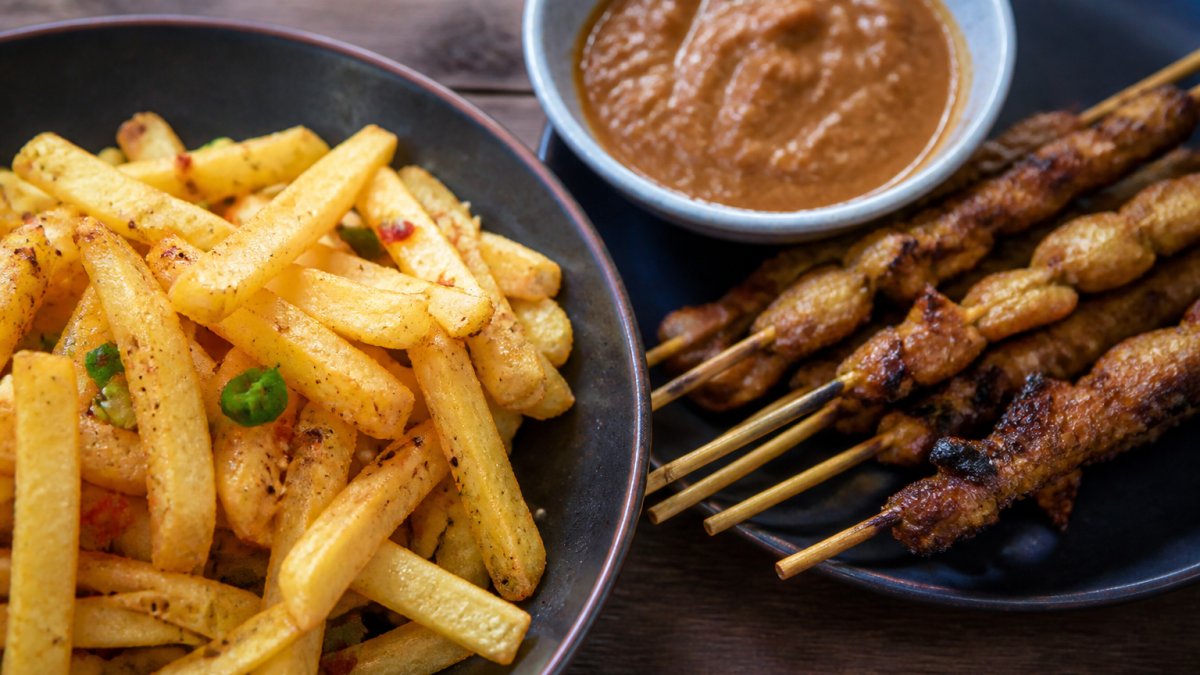 Close-up of crispy golden salt and chilli chips alongside a plate of Indonesian satay skewers with rich peanut sauce, showing the textural and colour contrast between the two styles