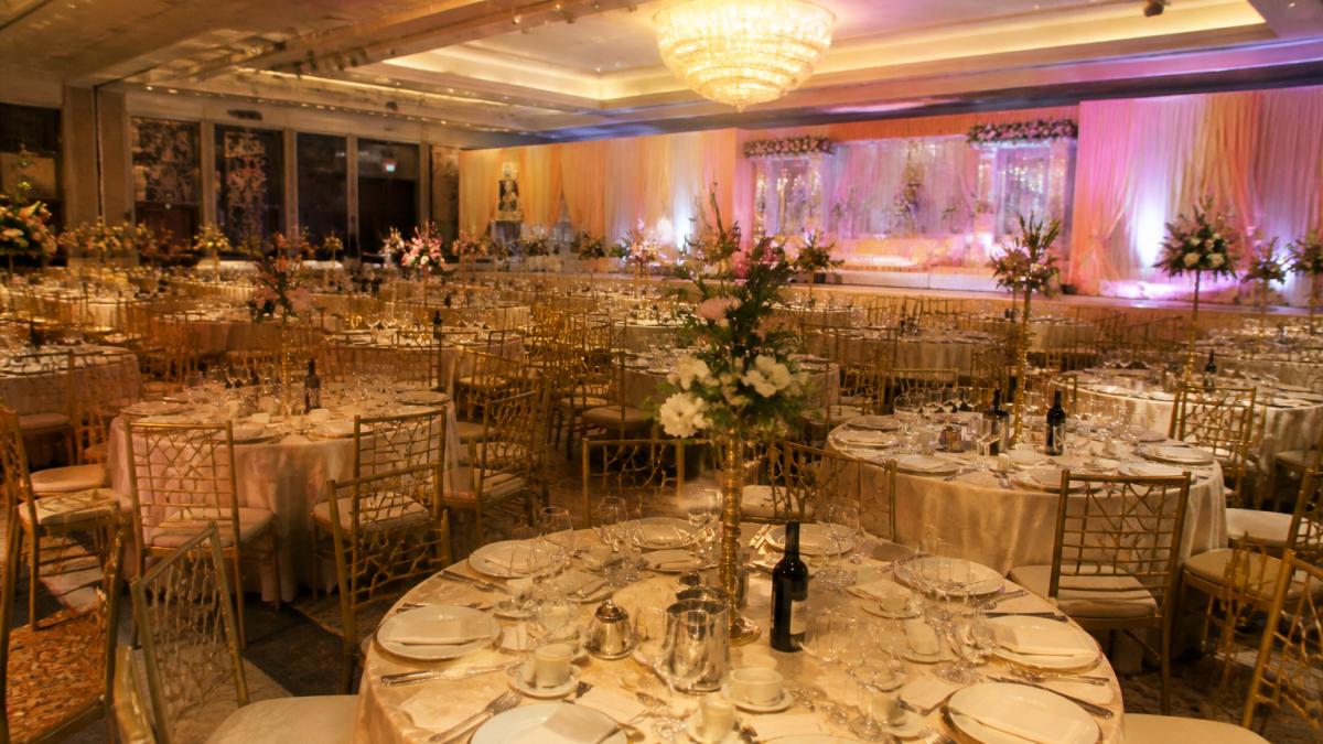 An elegantly decorated banqueting hall set for a large Asian wedding reception, with round tables, warm lighting and a stage area in the background