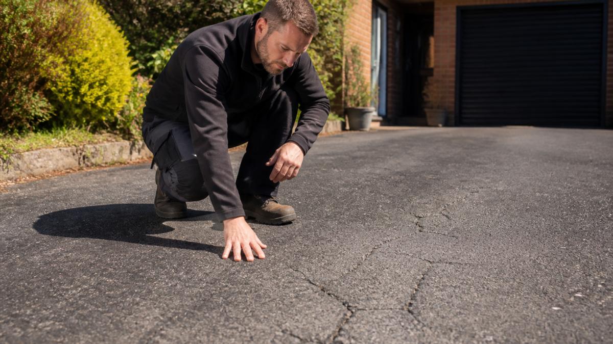 Professional inspecting an asphalt driveway surface for cracks and wear, with sunlight highlighting surface condition and texture