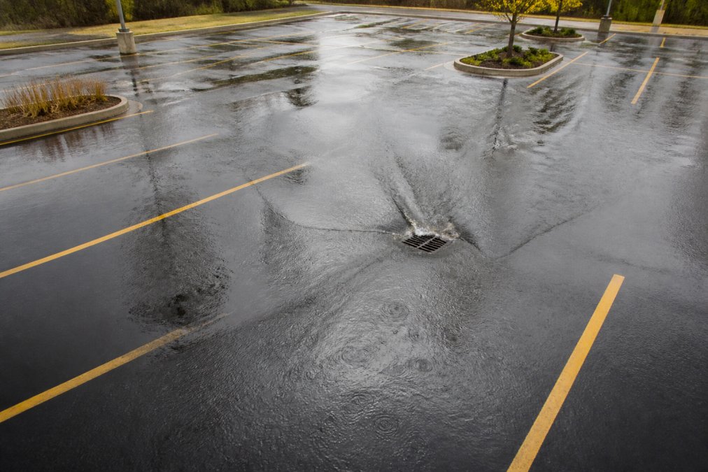 Aerial view of parking lot with visible water pooling and drainage issues during heavy rain, showing water management challenges on asphalt surface