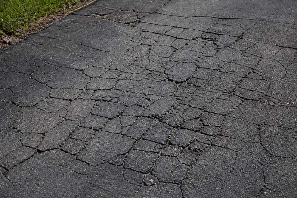 Close-up of severely deteriorated asphalt pavement with extensive alligator cracking patterns, dark asphalt showing interconnected fracture lines across multiple sections under bright daylight