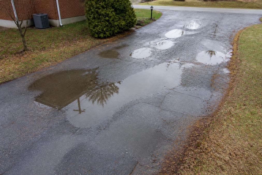 Aerial view of a residential driveway with visible water pooling in several low spots, showing standing water after recent rain, demonstrating poor drainage and moisture accumulation affecting pavement integrity