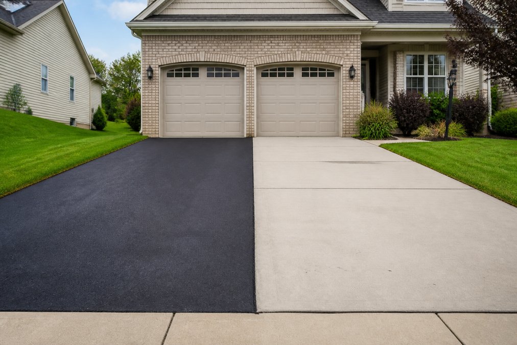 A residential driveway in a suburban neighborhood showing a clean black asphalt surface next to a light gray concrete driveway, with a two-car garage and green lawn in the background