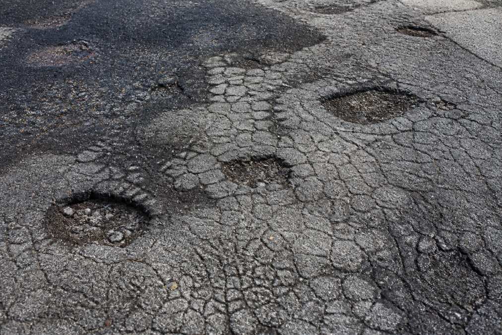 Close-up of asphalt driveway surface showing alligator cracking pattern, multiple potholes, and dark staining in natural daylight with shadow detail