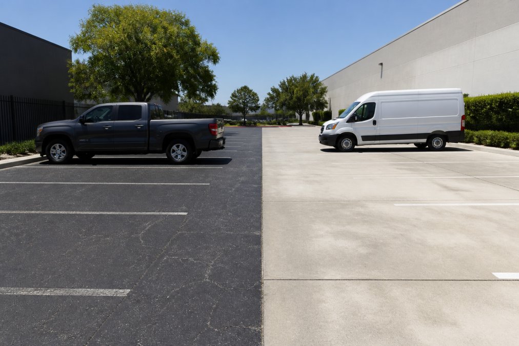 A commercial parking lot split down the middle, one half dark asphalt with faded white parking lines and a pickup truck parked on it, the other half light gray concrete with a delivery van, bright midday sun casting sharp shadows
