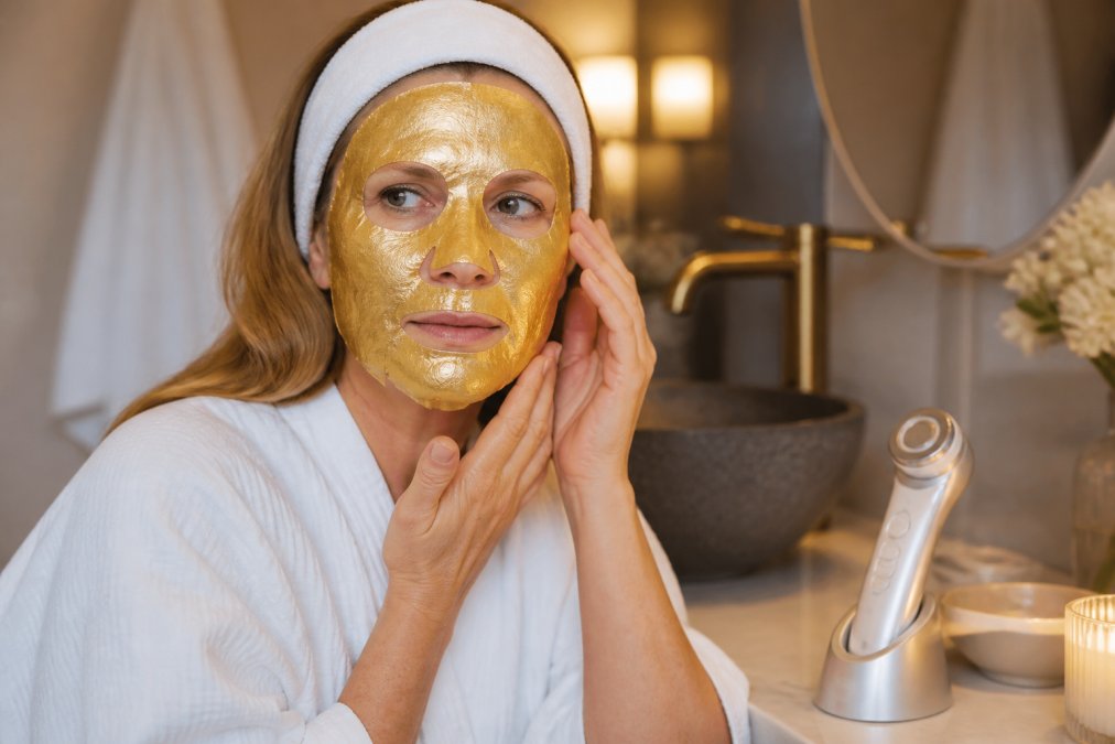 A woman in her 40s with glowing skin applying a gold collagen face mask at home while wearing a spa headband, with the EMS facial tool on the counter beside her, soft bathroom lighting