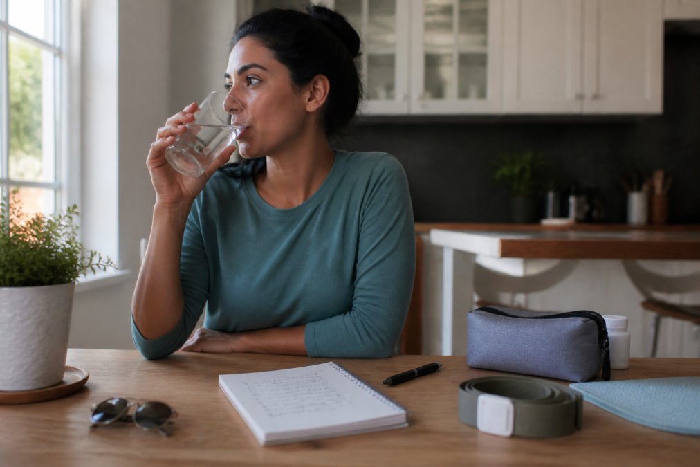 A person drinking a large glass of water at a bright kitchen table beside a notepad with a handwritten checklist, preparing calmly for an at-home medical appointment in morning light