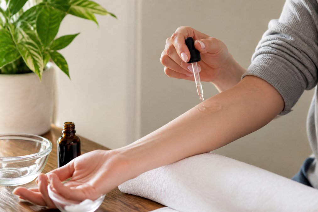 A person applying a small drop of clear serum from a glass dropper onto their inner forearm near the elbow, clean skin visible, soft natural window light