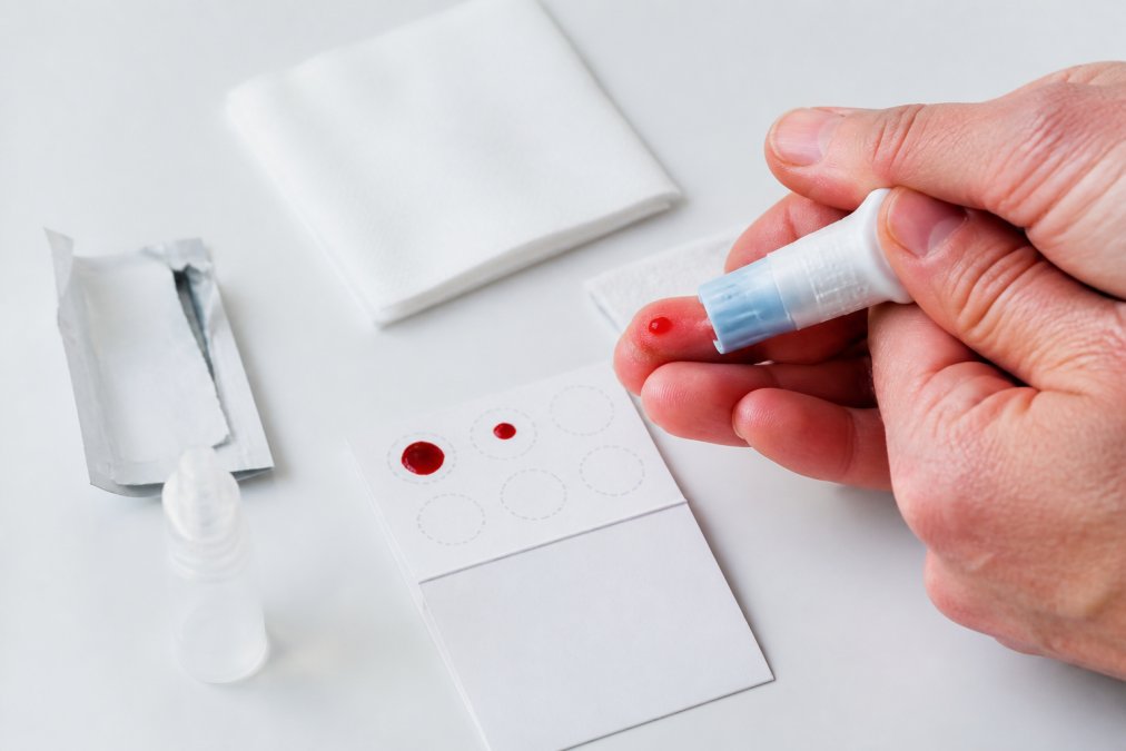 Close-up of a person's fingertip being pricked with a sterile lancet, with drops of blood falling onto a dried blood spot collection card placed on a clean white surface