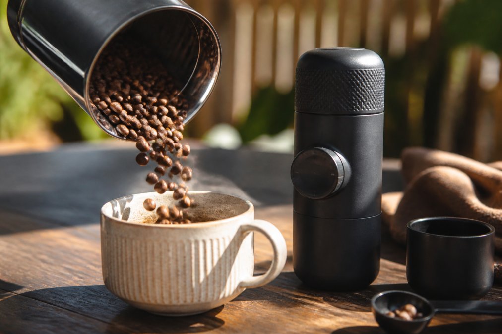 Friskristede kaffebønner bliver hældt fra en risteri-beholder ind i en elegant kaffekop, med hele bønner synlige i varmt naturlys, og en håndholdt rejse-espressomaskine placeret ved siden af