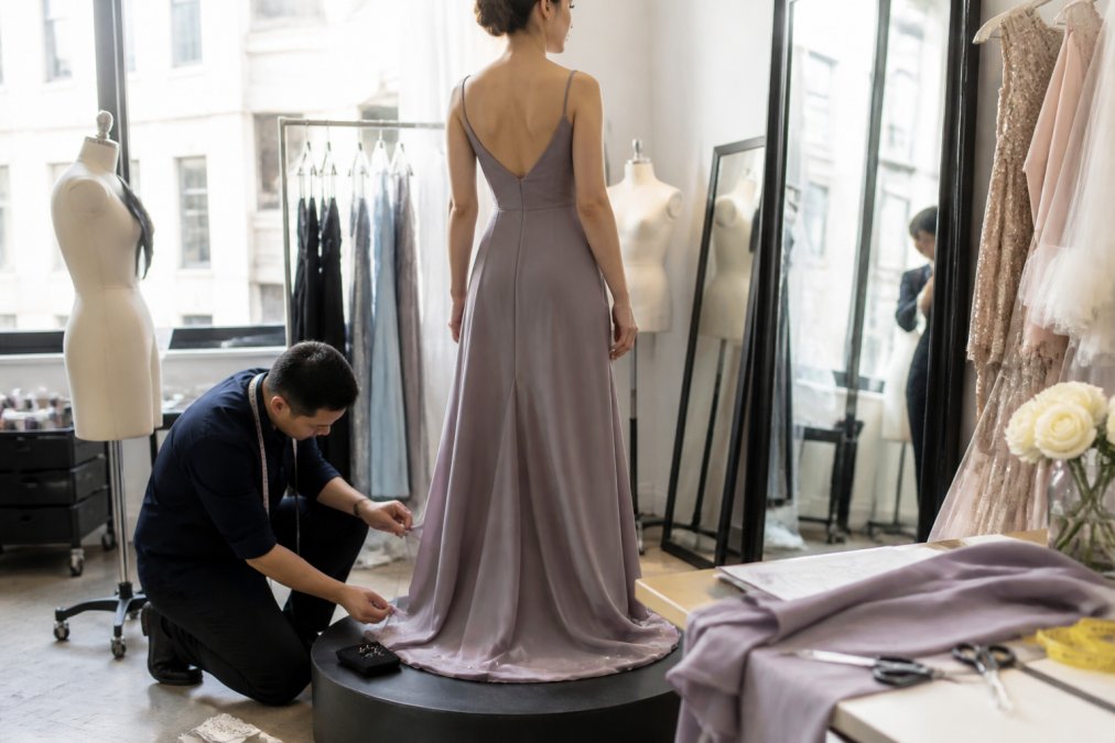 Professional tailor adjusting the hem and silhouette of an evening gown on a client standing on a fitting platform, using pins and chalk to mark alterations in a bright, well-lit atelier studio
