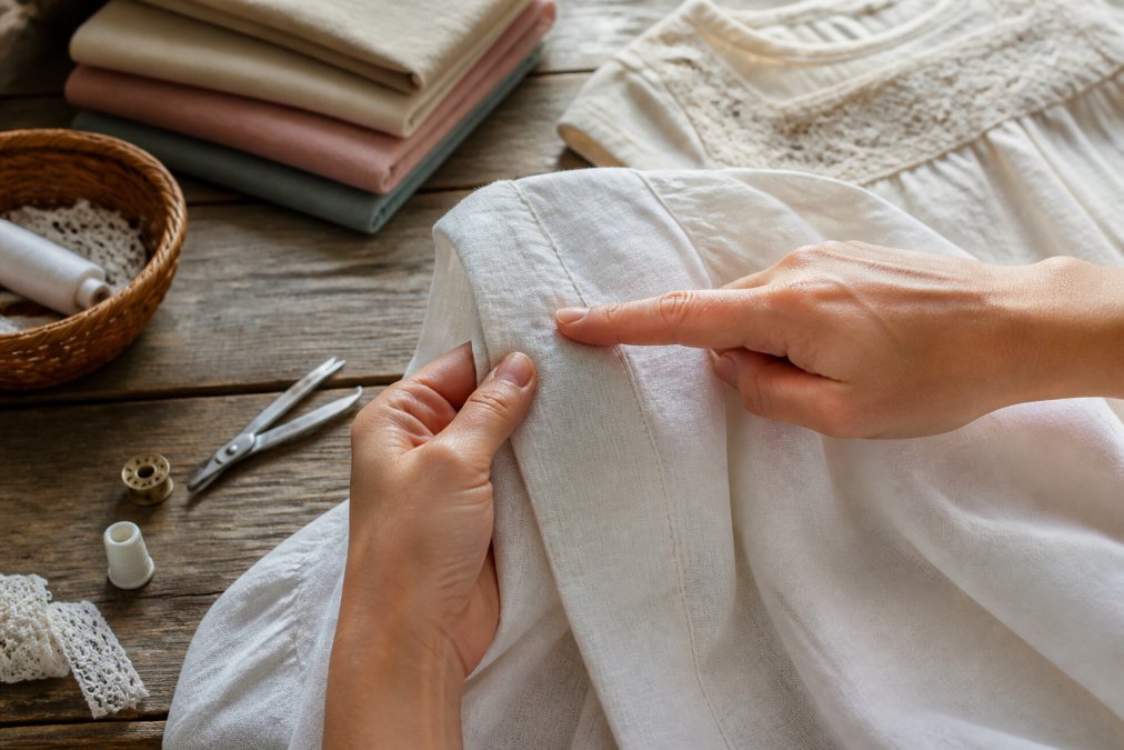 Close-up of hands carefully examining seam stitching on a white linen garment, fingers tracing the precise needle work along a shoulder seam in natural daylight
