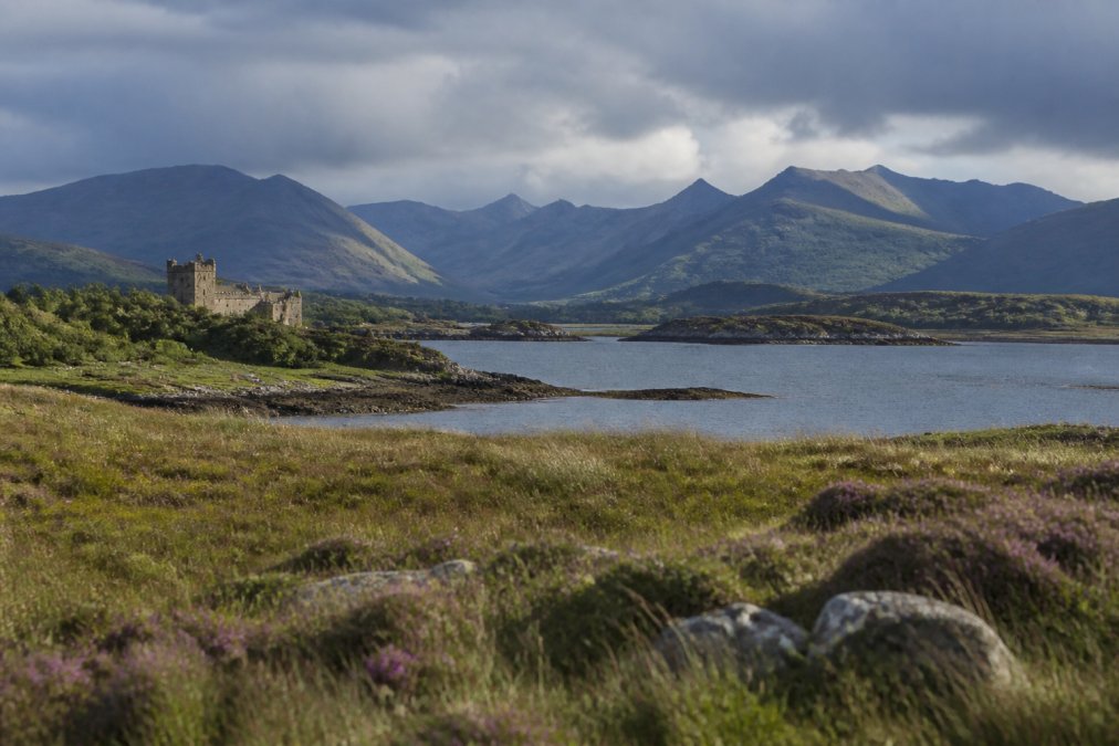 Dramatic Highland landscape showing Dunvegan Castle visible in the distance across moorland, with coastal views and Cuillin mountains on the horizon under moody Scottish sky