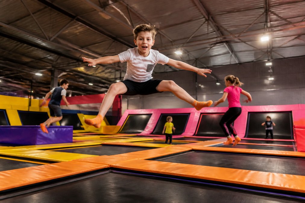 Dzieci skaczące na trampolinach w nowoczesnej hali, uśmiechnięte i aktywne, z bezpieczną infrastrukturą widoczną w tle, jasne oświetlenie