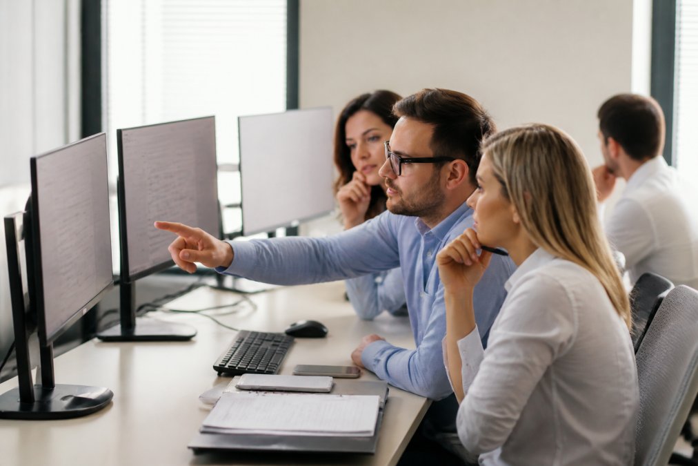 Recruitment team reviewing candidate profiles on multiple computer screens in a modern office environment, with one professional pointing at a monitor displaying candidate information and assessment scores