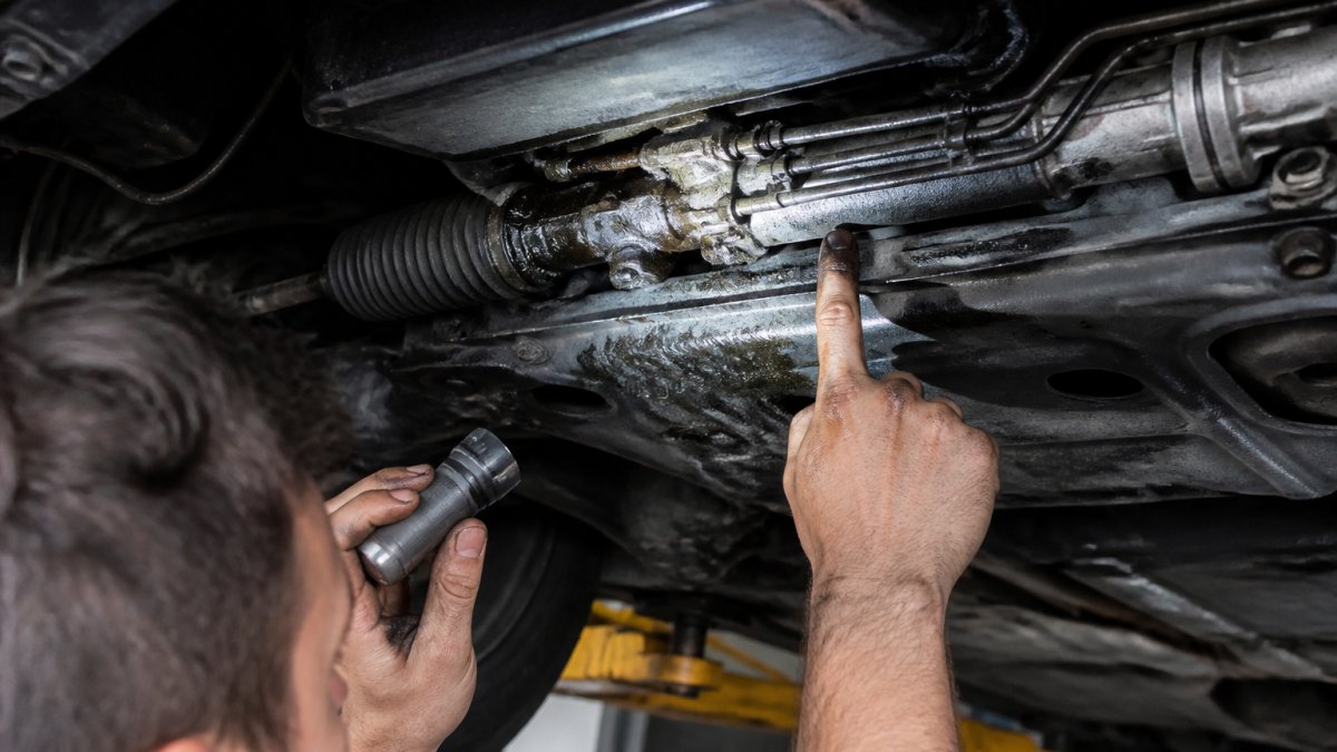Close-up of hands performing an under-chassis inspection on an Audi A5, with a flashlight illuminating the steering rack and hydraulic hoses, showing fluid residue on corroded pipework and connection points