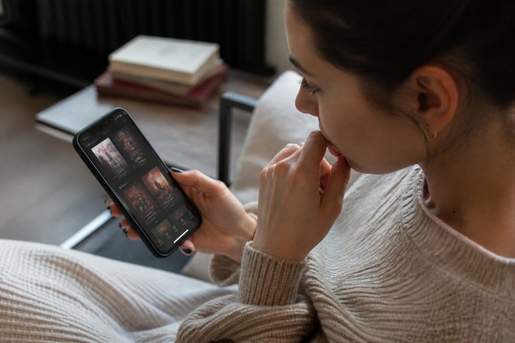 Close-up of hands holding a smartphone displaying a podcast or audio app interface with content thumbnails visible, the person's face showing thoughtful consideration while browsing