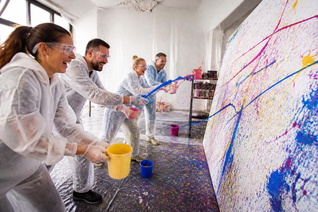 Eine Gruppe von Erwachsenen in Schutzkitteln und mit Schutzbrillen wirft Farbe auf eine große Leinwand in einem hellen Atelierraum, lachend und voller Energie, mit Farbspritzern auf dem Boden und an den Wänden