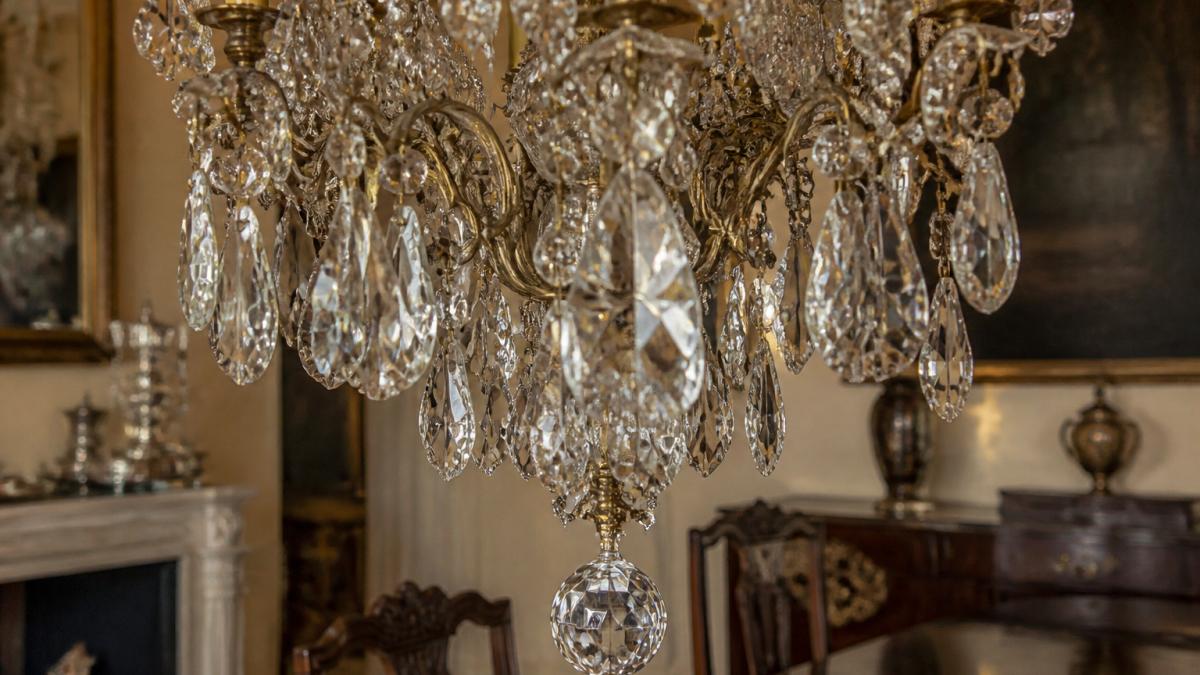 Close-up of an ornate crystal chandelier with intricate cut glass prisms and brass fittings, hanging in an elegant dining room with soft golden light reflecting through the crystals