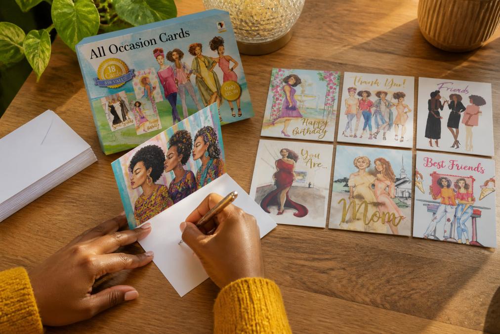 Close-up of hands writing a thoughtful message on a premium greeting card featuring vibrant African-inspired geometric patterns and illustrations of Black women, with matching white envelopes and additional cards arranged on a warm wooden desk with natural afternoon light