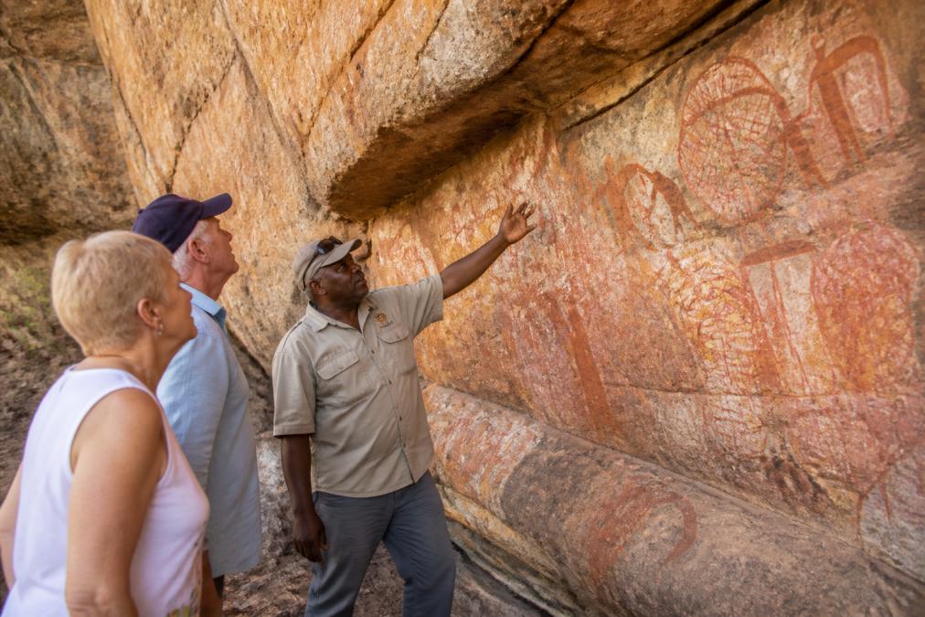 Close-up of ancient ochre rock art on canyon wall with weathered patterns and symbols, Indigenous guide gesturing to explain the artistic and cultural significance to visitors