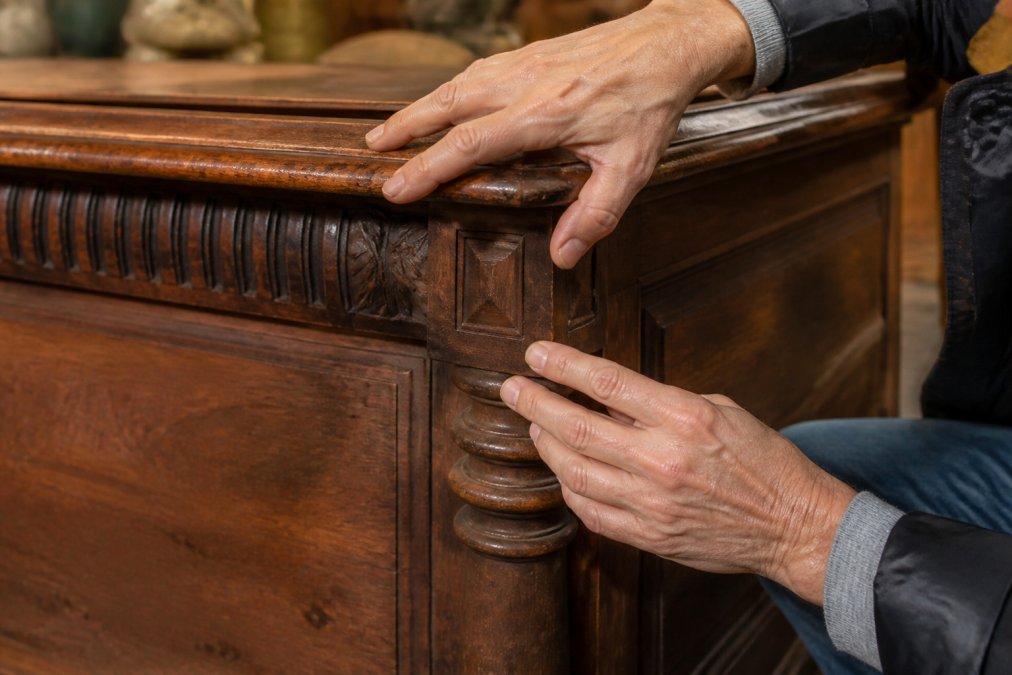 Close-up of hands examining antique wood joinery and hand-carved details on period furniture, showing grain patterns and construction techniques under natural light