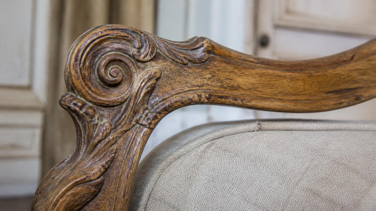 Close-up of hand-carved wooden details on an 18th-century French provincial chair showing intricate rocaille scrollwork, wood grain patterns, and natural patina from age