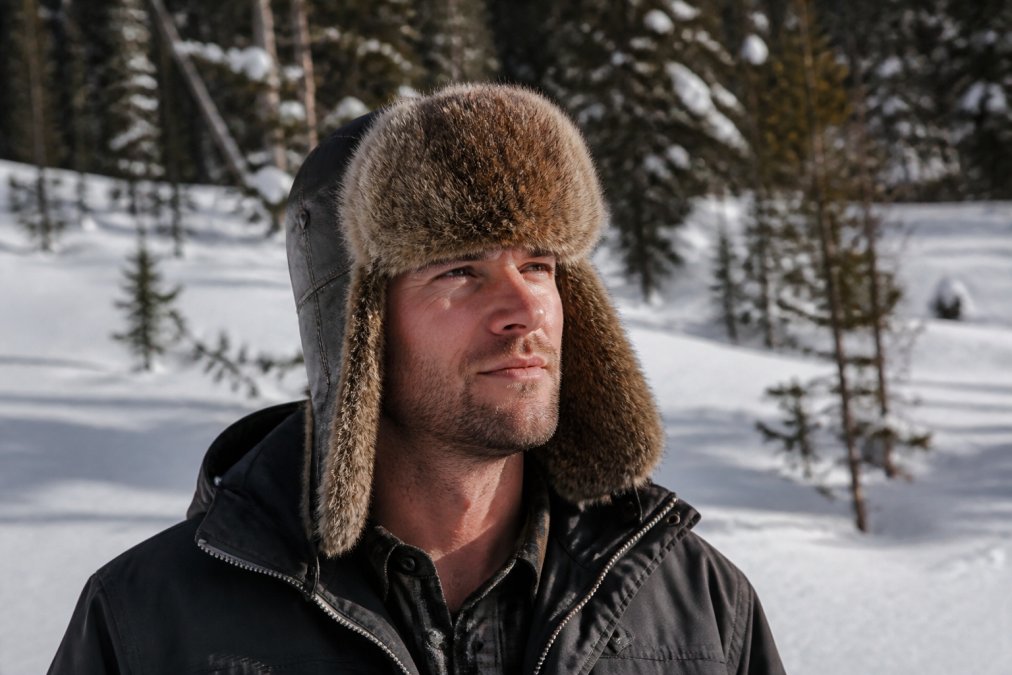 Man wearing a premium fur aviator hat outdoors in winter conditions, showing natural fur texture, properly fitted ear flaps, and the hat's silhouette against snow-covered landscape with natural daylight