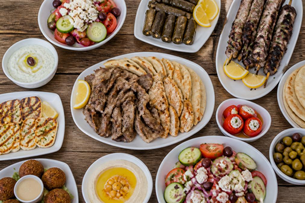 Overhead view of Mediterranean catering spread with fresh mezze platters, grilled lamb and chicken, colorful Greek salads with feta, olives, and pita bread arranged on white ceramic serving dishes on a rustic wooden table