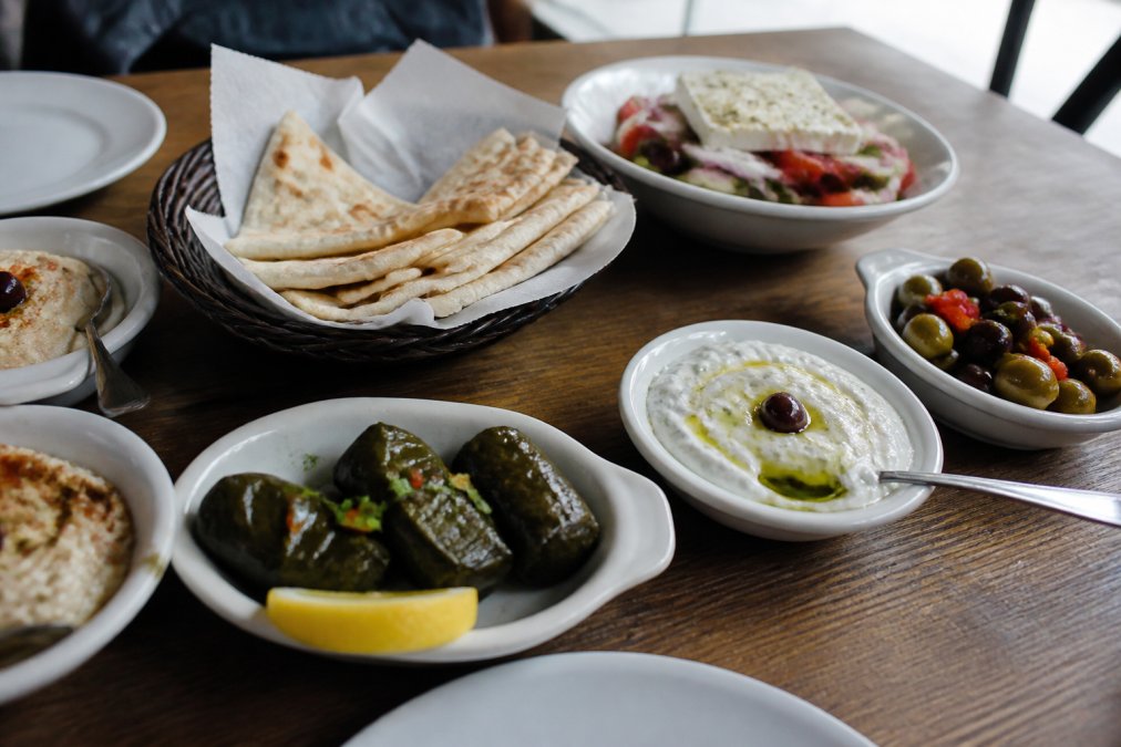 A wooden table filled with mezze plates including tzatziki, olives, dolmades, and warm pita bread, photographed in natural light at a restaurant