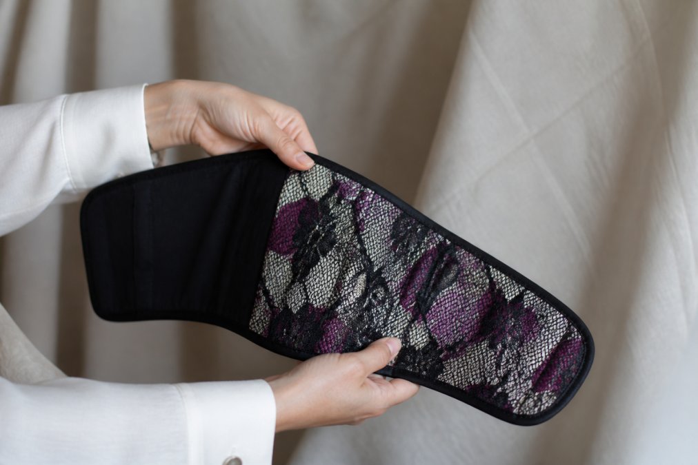 Close-up of hands holding a reversible ikat corset belt, showing the detailed ikat pattern layered with French lace on one side and the solid black reverse, displayed against a neutral background with soft natural lighting