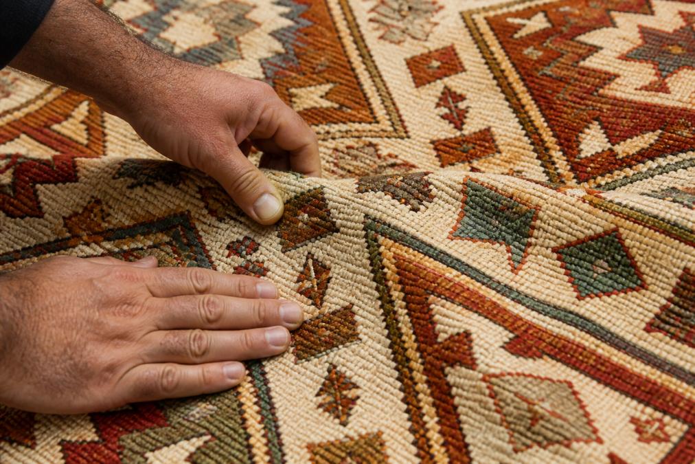 Close-up of hands examining the woven texture of an authentic kilim rug, showing intricate geometric patterns in natural wool with earth tones and tribal motifs, natural daylight highlighting the flatweave construction