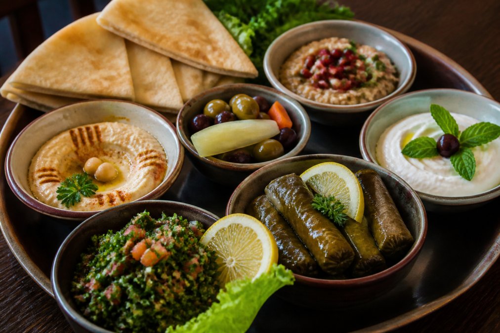 Overhead close-up of a beautifully arranged Lebanese mezze platter on a dark wood table, featuring hummus, tabbouleh, stuffed grape leaves, warm pita bread, and garnishes in small ceramic dishes under warm restaurant lighting