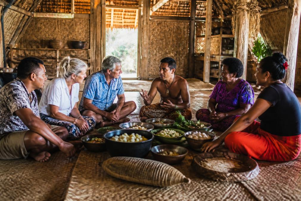 Local villagers and travellers sitting together in a traditional bure longhouse, sharing a meal and conversation in warm afternoon light, with woven walls and traditional cooking implements visible