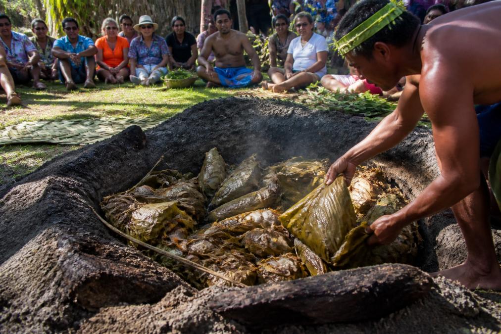 An earthen oven (umu) being opened to reveal traditional cooked food wrapped in leaves, with local people gathered around in a village setting under natural light